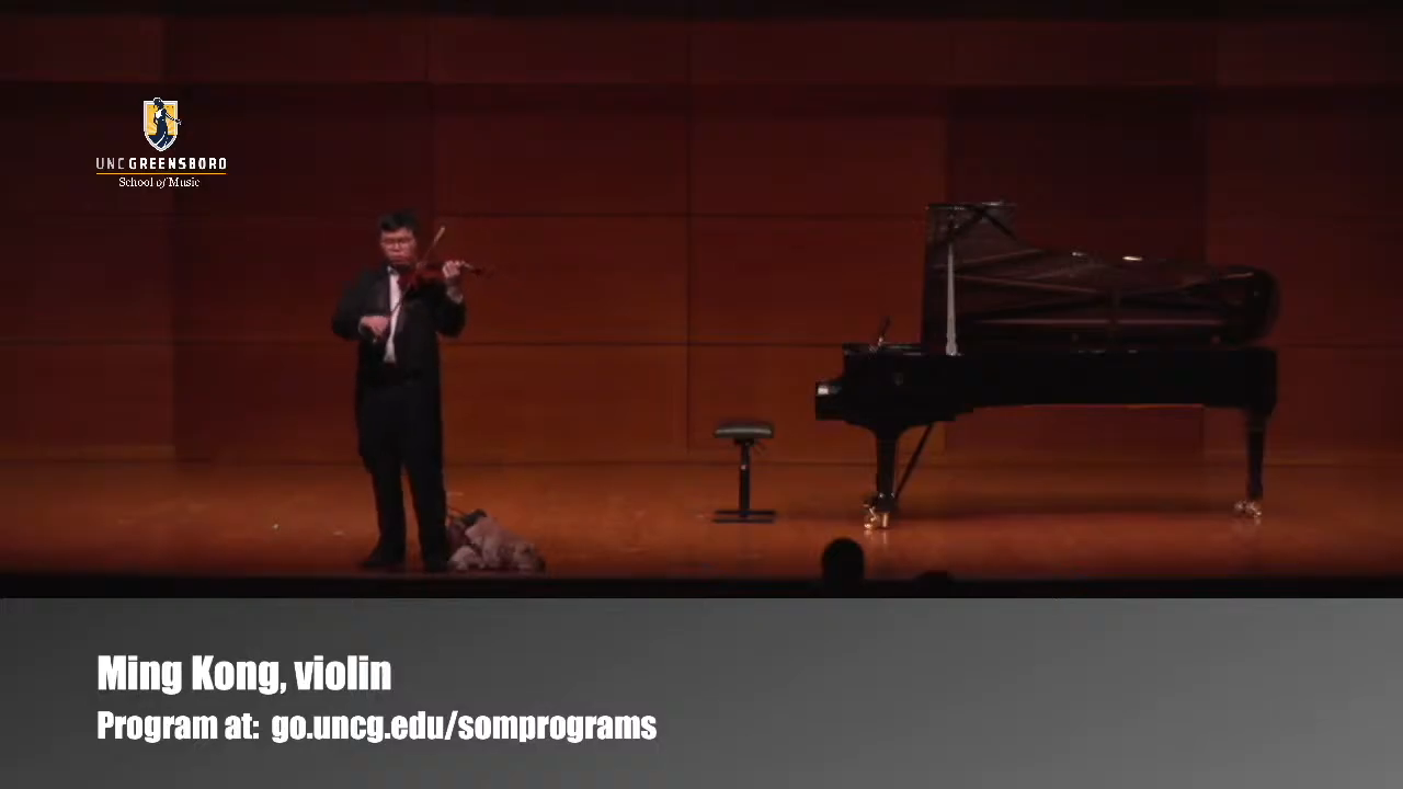 Ming Kong playing violin on the left of a UNCG concert stage, with a grand piano on the right; the video identifies him as Ming Kong, violin.